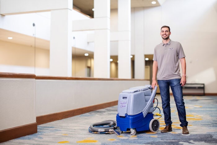 A man stands in a hallway holding the handle of a large carpet cleaning machine with a hose attachment on a patterned carpet floor.