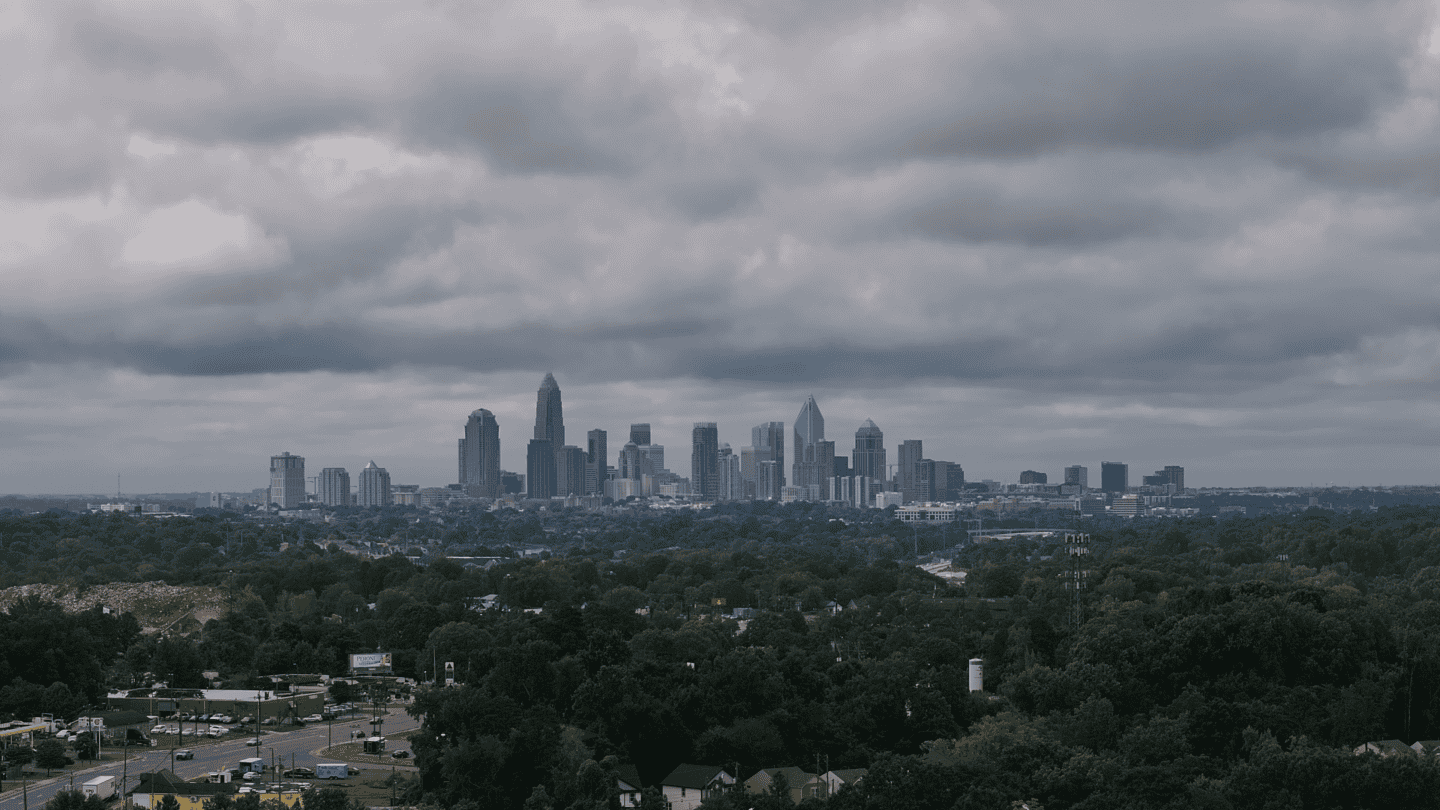 A city skyline with tall buildings under a cloudy sky, surrounded by trees and residential areas in the foreground.