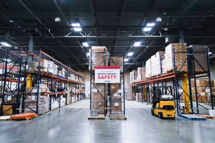Interior of a warehouse with tall shelves of stacked boxes, a forklift, and a sign reading "Service Through Safety" in the central aisle.