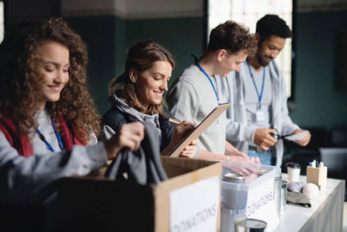 Four people stand at a donations table, sorting and organizing items; one writes on a clipboard while others handle clothing and supplies.