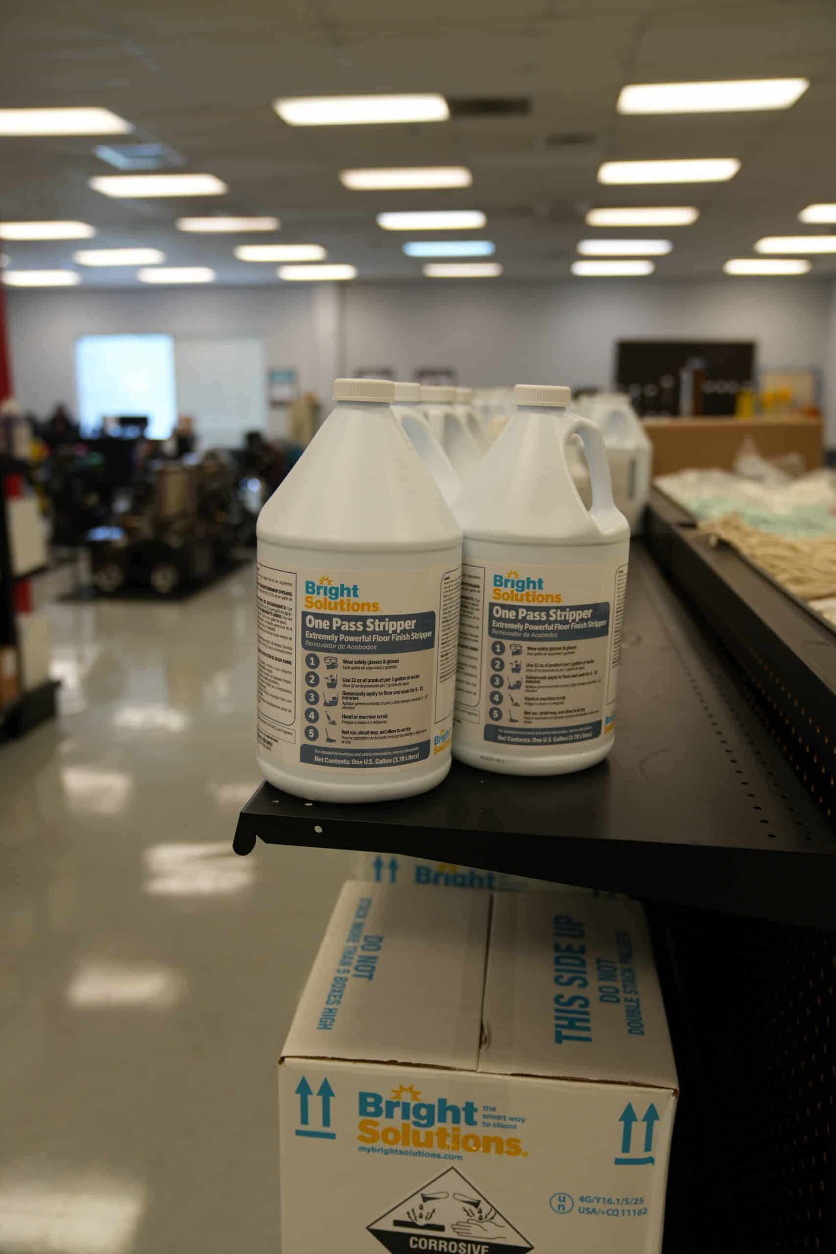 Three gallon-sized containers of Bright Solutions One Pass Stripper are displayed on a black shelf in a well-lit commercial or warehouse setting.