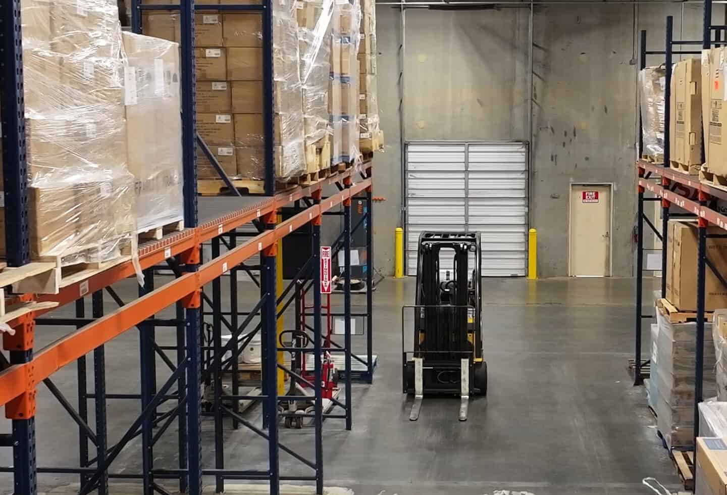 A forklift is parked in the aisle of a warehouse with tall shelves stacked with pallets and boxes on both sides.