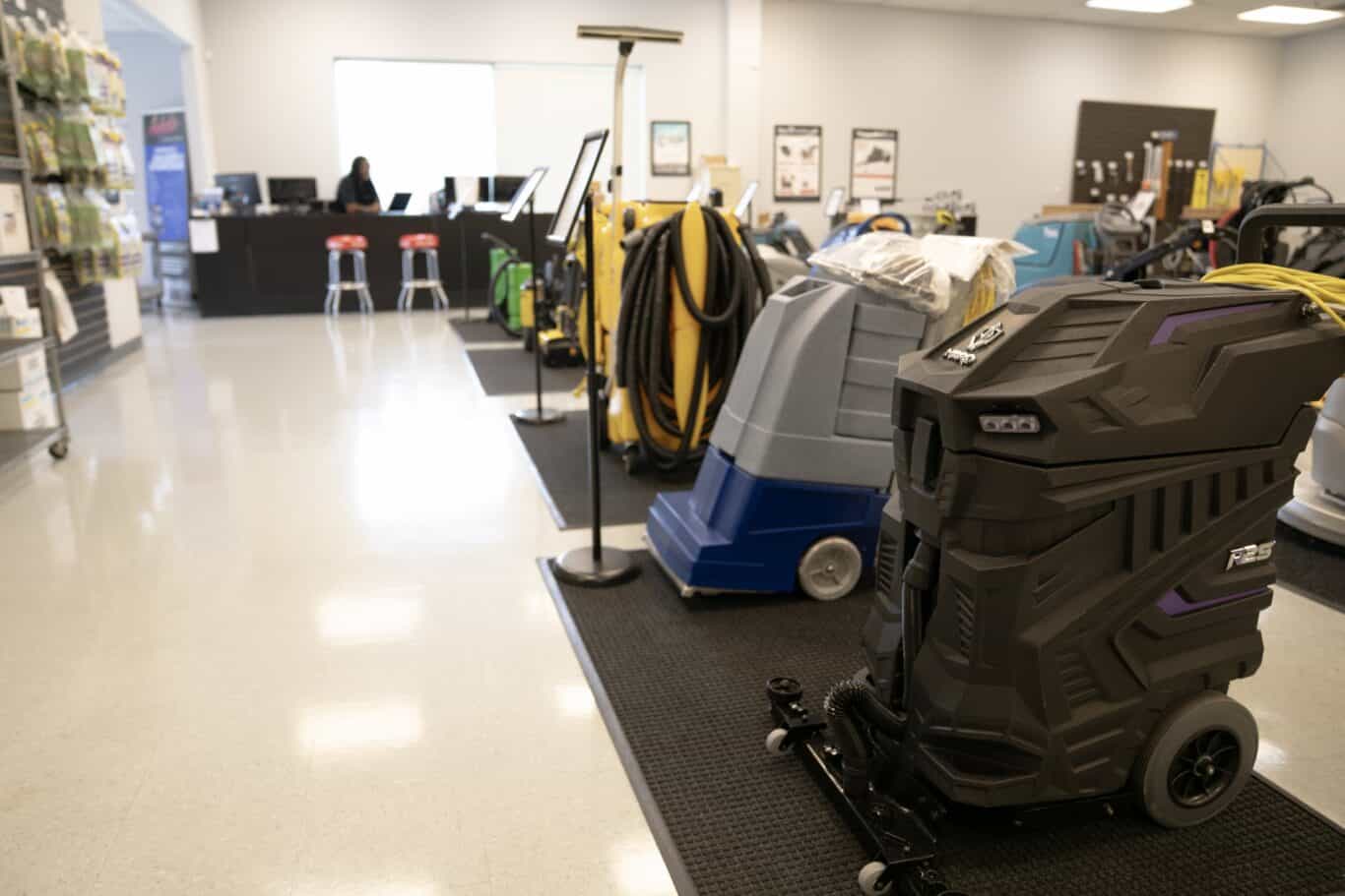 A showroom displays various commercial cleaning machines on mats; a person sits at a desk in the background near a bright window.