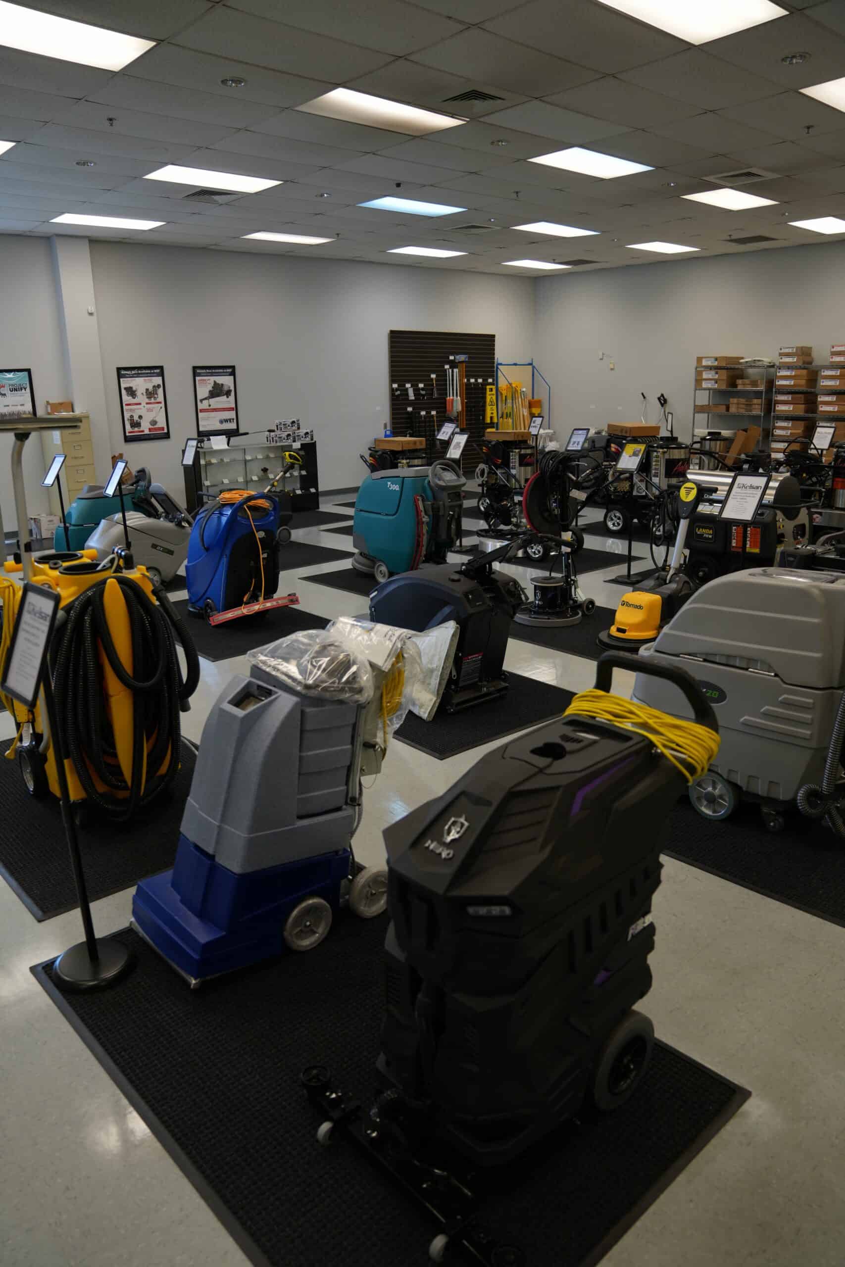 A showroom displays various commercial floor cleaning machines and equipment, organized on mats under bright overhead lights.