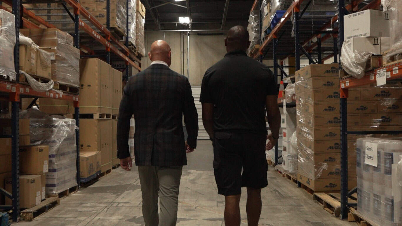 Two men walk down an aisle in a warehouse, surrounded by shelves of stacked boxes and pallets.