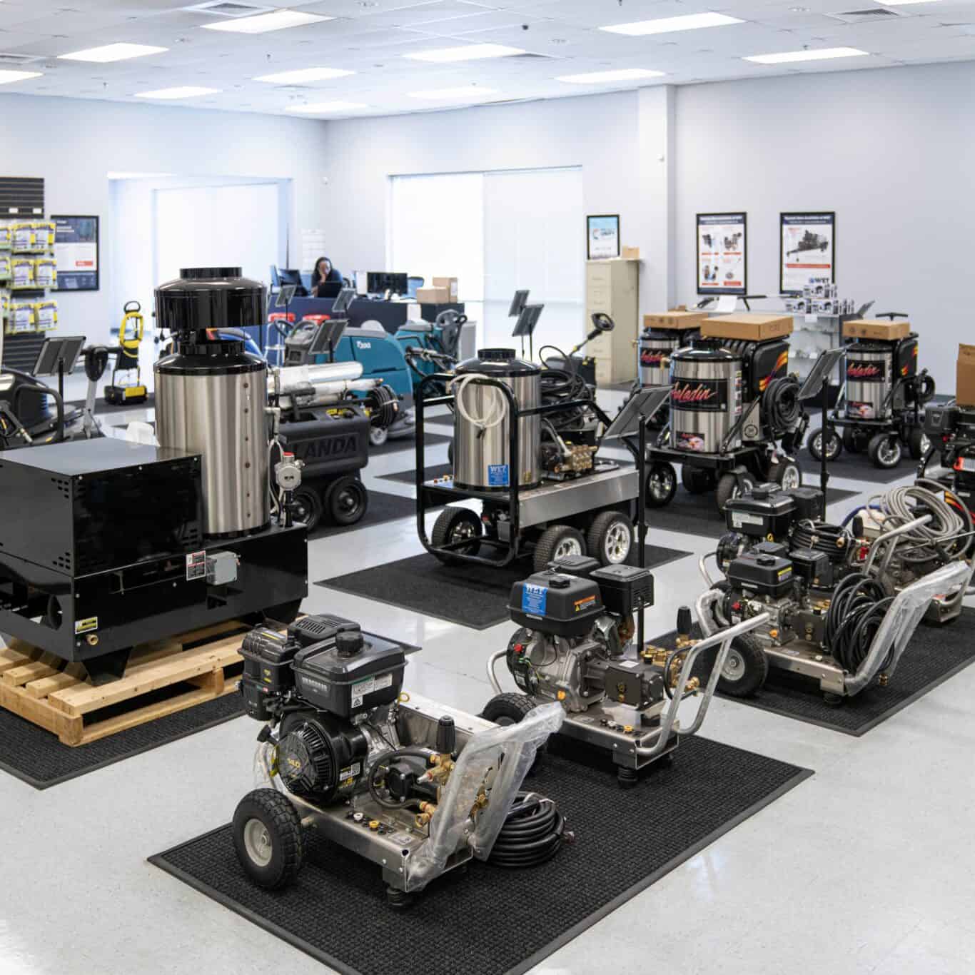 Showroom displaying multiple industrial floor cleaning and pressure washing machines neatly arranged on mats, with a person working at a desk in the background.