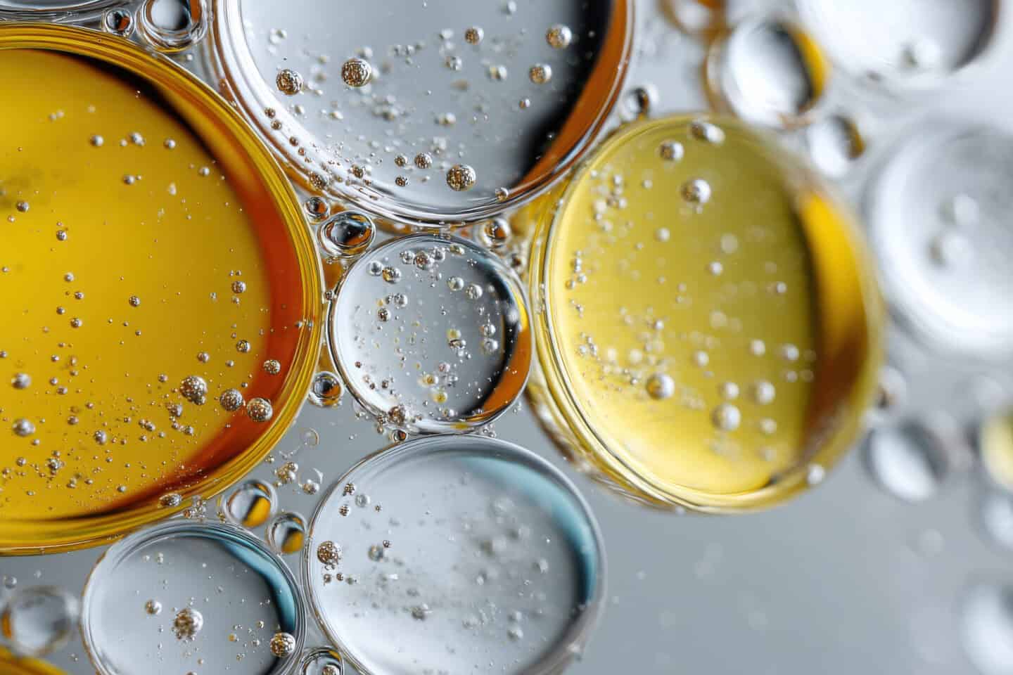 Close-up image of oil and water droplets of various sizes, creating circular patterns with reflections and bubbles against a neutral background.